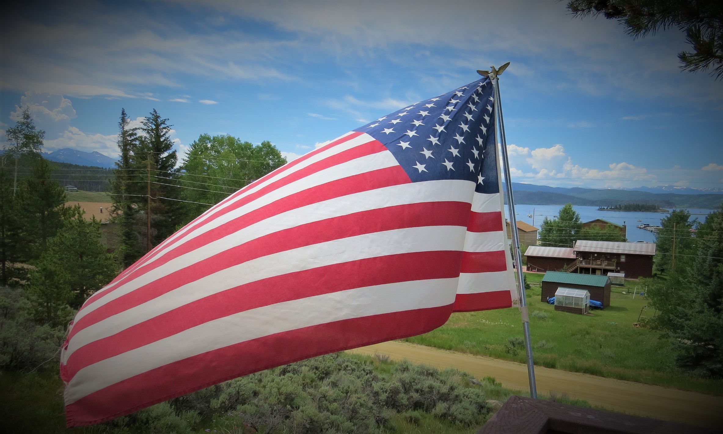 U.S. Flag waving in the breeze with mountain and lake scenic background on the Fourth of July during the COVID-19 pandemic. The accompanying article discusses the idea of potential in healing after trauma, in this case a shared, collective trauma.