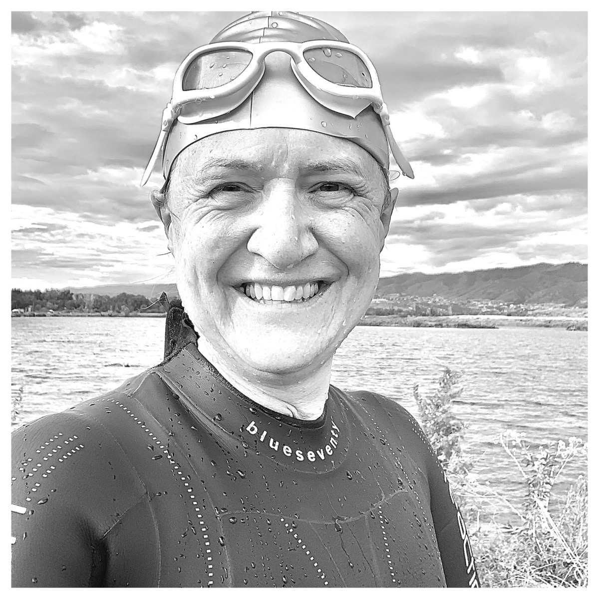 Black and white photo of the author in wetsuit, swimsuit and goggles standing next to a body of water, smiling.