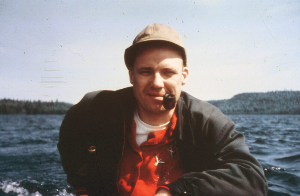 Photo of author's dad in a boat in the 1960s with dark jacket, red short, tan cap and a pipe in his mouth.